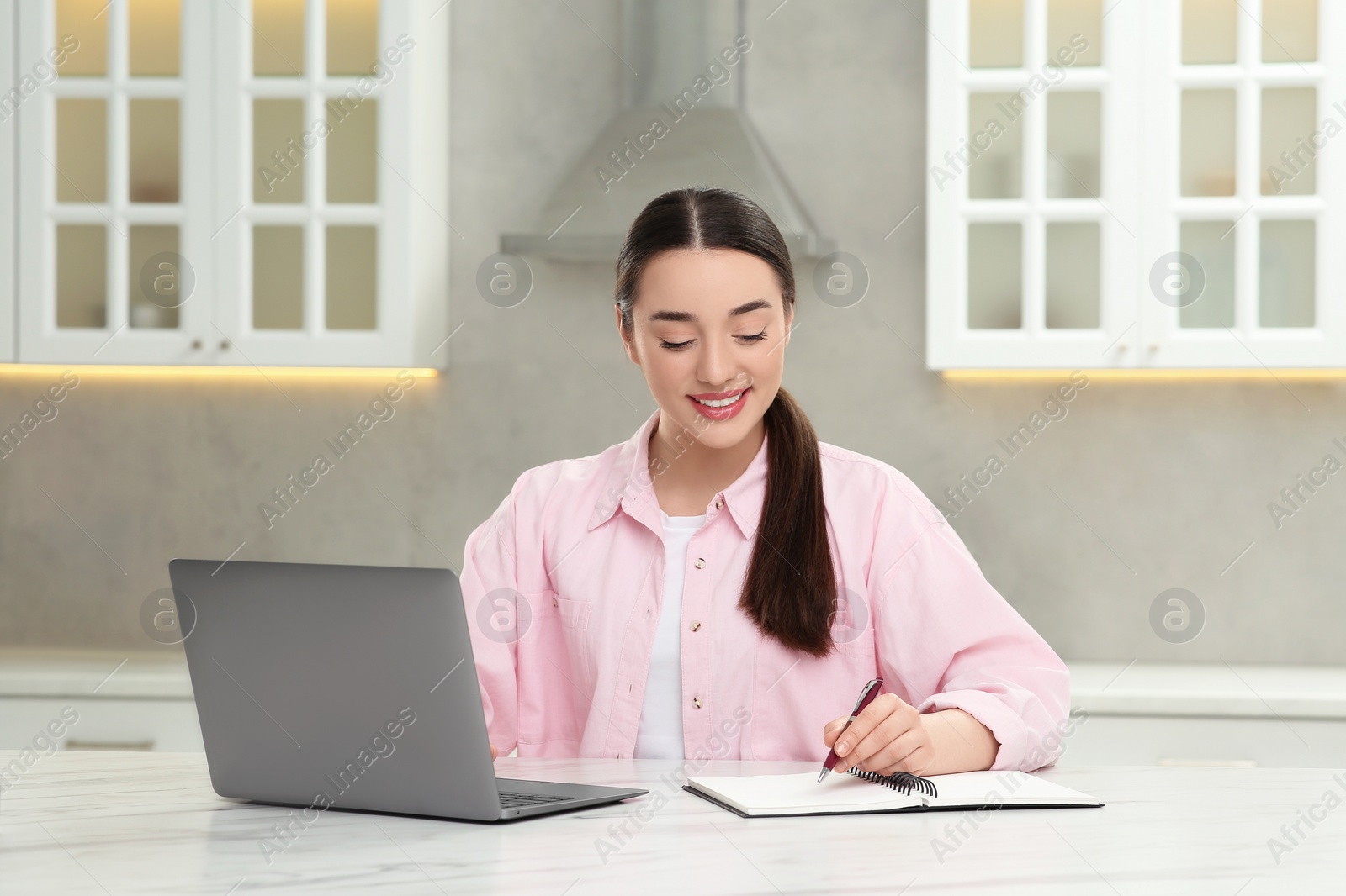 Photo of Woman writing something in notebook while using laptop at white table in kitchen