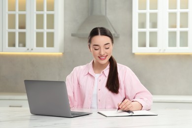 Woman writing something in notebook while using laptop at white table in kitchen