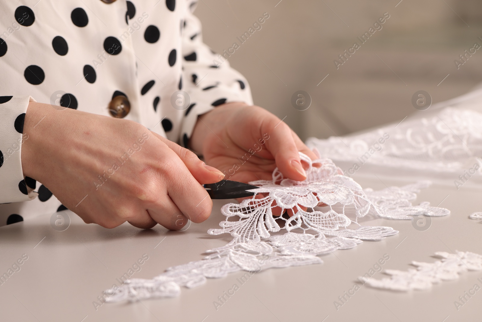 Photo of Dressmaker cutting beautiful white lace at table in atelier, closeup