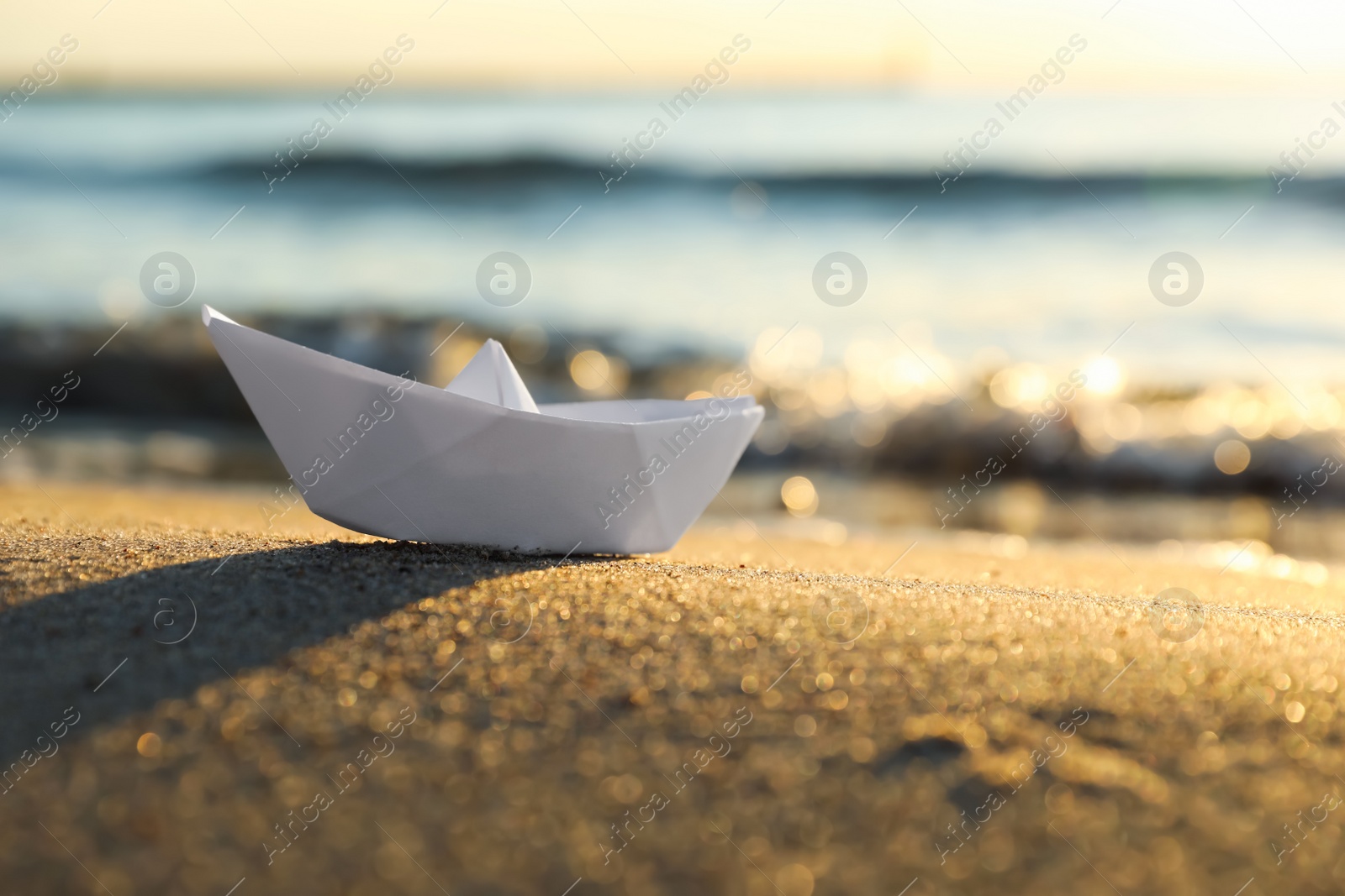 Photo of White paper boat on sand near sea at sunset, closeup. Space for text