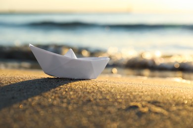 Photo of White paper boat on sand near sea at sunset, closeup. Space for text