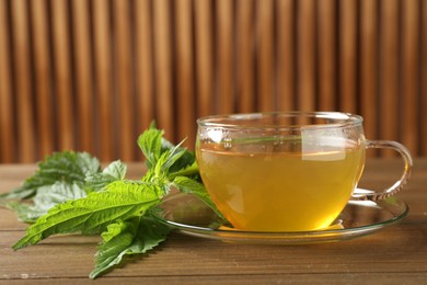 Photo of Glass cup of aromatic nettle tea and green leaves on wooden table, closeup