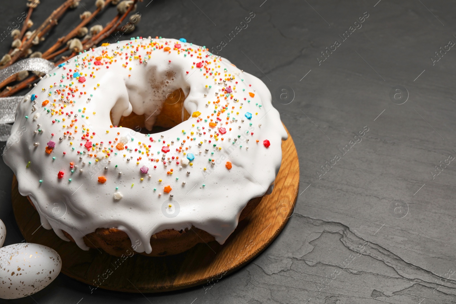 Photo of Delicious Easter cake decorated with sprinkles near egg and willow branches on black textured table, closeup. Space for text