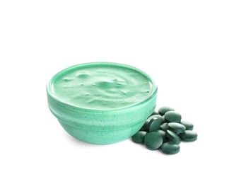 Freshly made spirulina facial mask in bowl and pills on white background