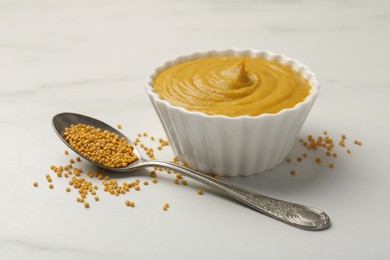 Photo of Tasty mustard sauce in bowl and spoon with dry seeds on white marble table