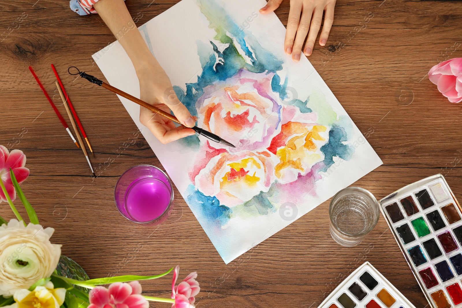 Photo of Woman painting flowers with watercolor at wooden table, top view. Creative artwork