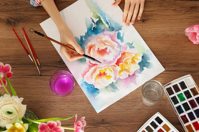 Photo of Woman painting flowers with watercolor at wooden table, top view. Creative artwork