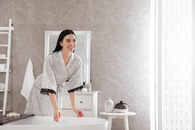 Photo of Beautiful young woman in silk robe near bathtub at home