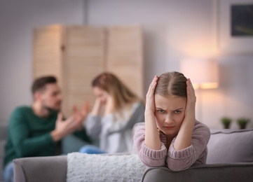 Little unhappy girl sitting in armchair while parents arguing at home