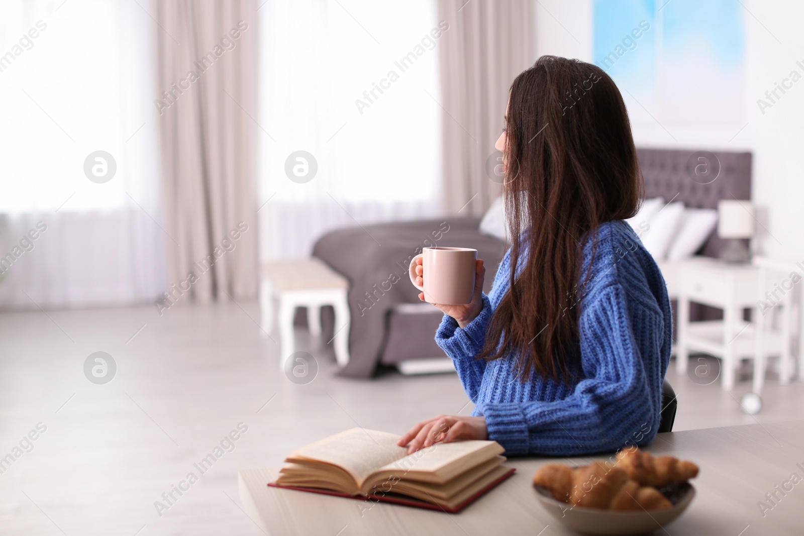 Photo of Young woman drinking coffee and reading book at table indoors, space for text. Winter season