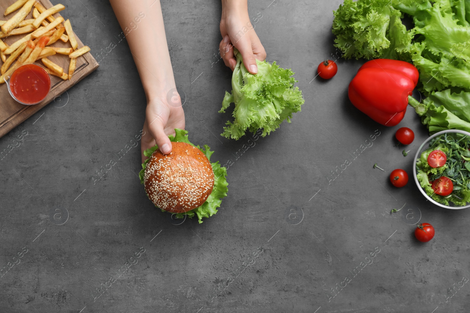 Photo of Top view of woman choosing between vegetables and burger with French fries at black table, closeup