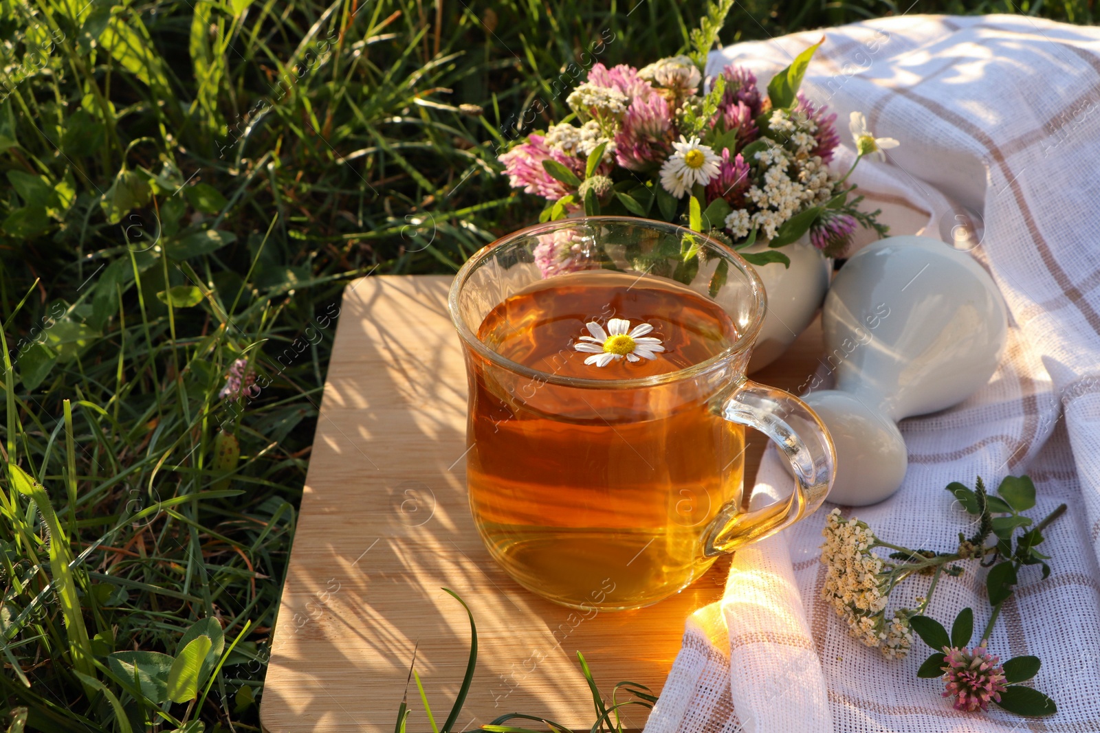 Photo of Cup of aromatic herbal tea, pestle and ceramic mortar with different wildflowers on green grass outdoors. Space for text