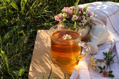 Photo of Cup of aromatic herbal tea, pestle and ceramic mortar with different wildflowers on green grass outdoors. Space for text