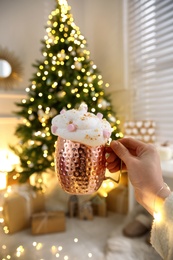 Photo of Woman holding cup of delicious drink with whipped cream and marshmallows near Christmas tree indoors, closeup