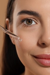 Beautiful young woman applying serum onto her face on beige background, closeup