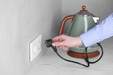 Photo of Woman plugging electric kettle into power socket at white table indoors, closeup