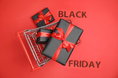 Photo of Small shopping cart with gift boxes and phrase Black Friday on red background, top view