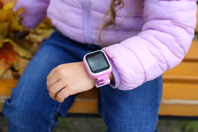 Photo of Little girl with stylish smart watch on bench outdoors, closeup
