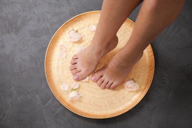 Woman soaking her feet in plate with water and roses on grey background, top view. Spa treatment