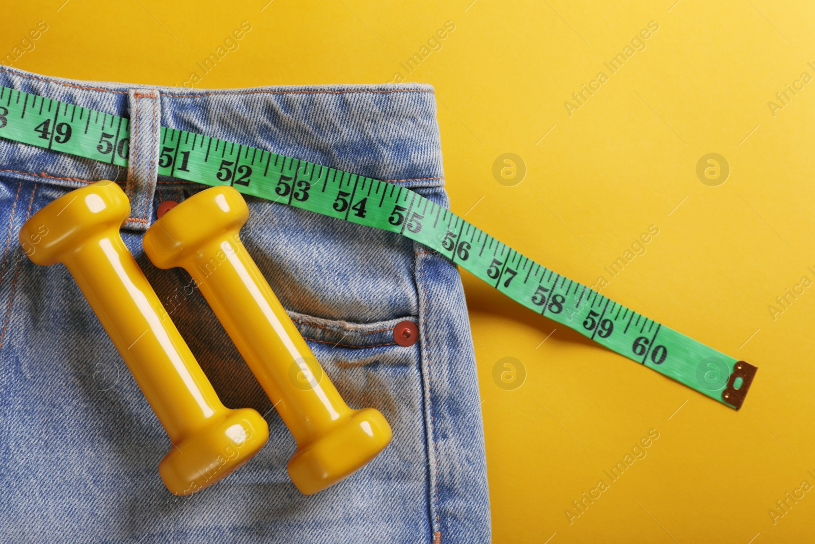 Photo of Jeans with measuring tape and dumbbells on yellow background, top view. Weight loss concept