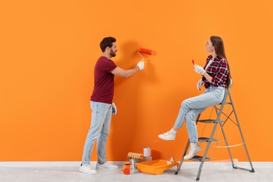 Photo of Man painting orange wall and woman sitting on folding ladder indoors. Interior design