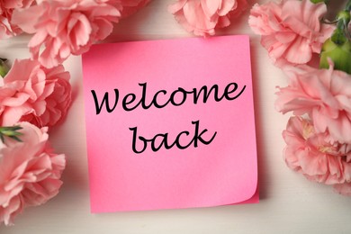 Image of Paper note with phrase Welcome Back and flowers on light table, flat lay