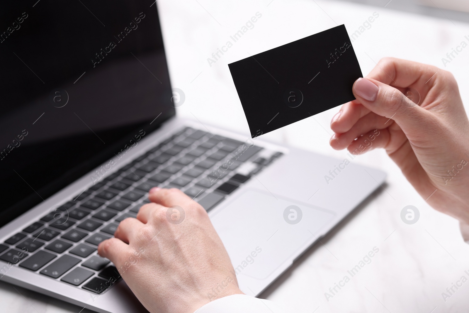 Photo of Woman with laptop holding blank business card at white table, closeup. Space for text