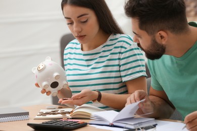 Young couple with piggy bank counting money at wooden table indoors