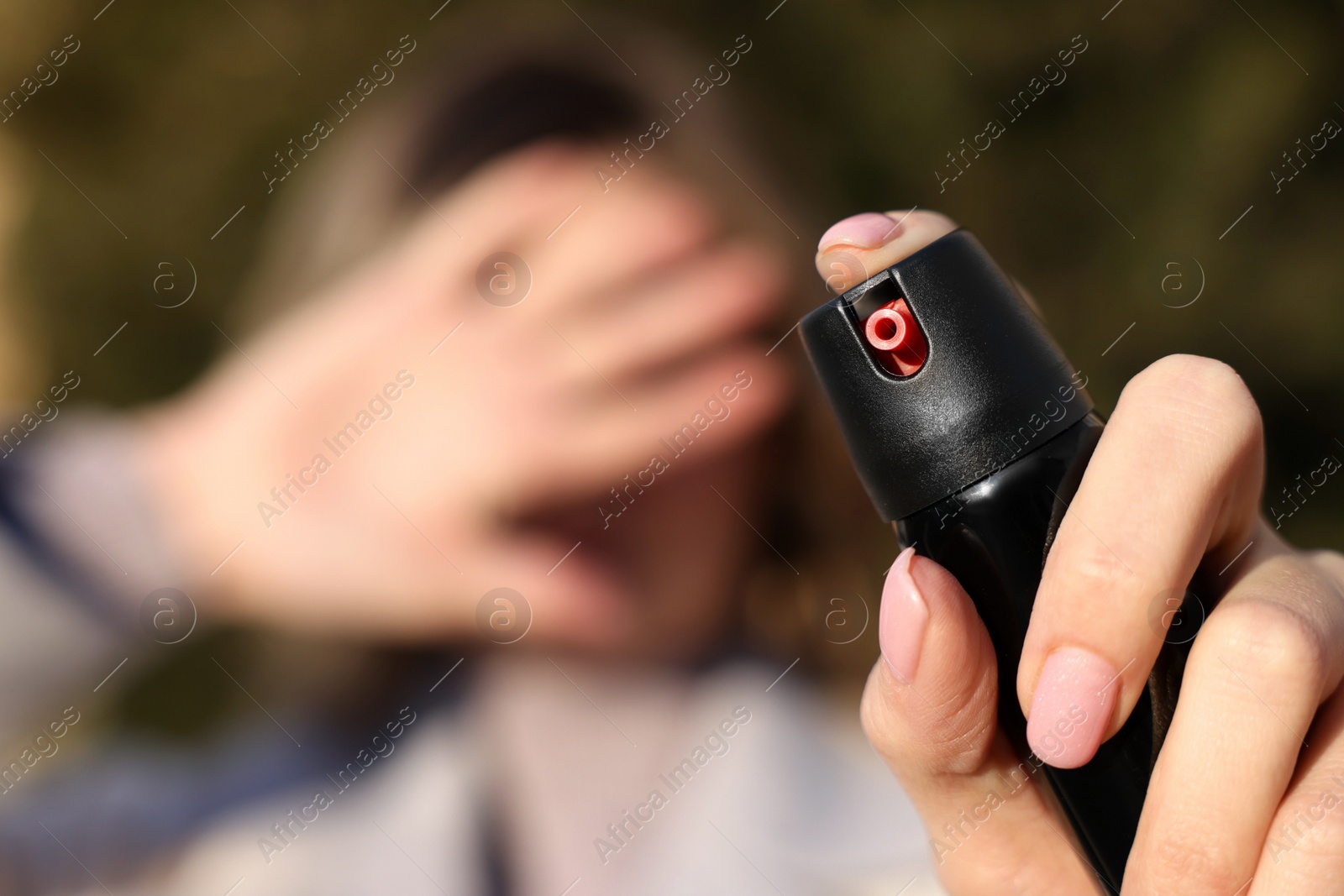 Photo of Young woman covering eyes with hand and using pepper spray outdoors, focus on canister