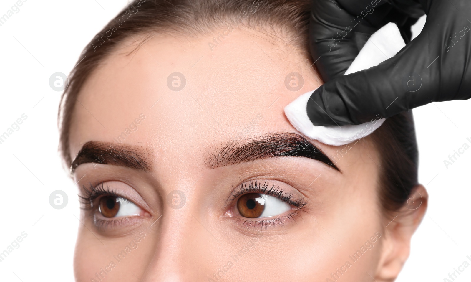 Photo of Beautician wiping tint from woman's eyebrows on white background, closeup