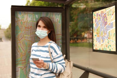 Image of Young woman in medical face mask waiting for public transport at bus stop