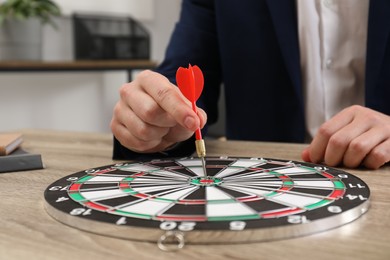 Photo of Business targeting concept. Man with dart aiming at dartboard at wooden table indoors, closeup