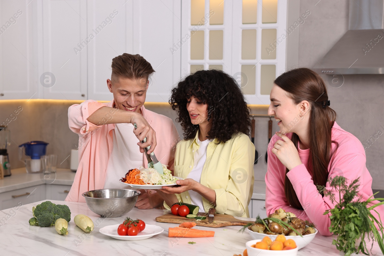 Photo of Friends cooking healthy vegetarian meal at white marble table in kitchen