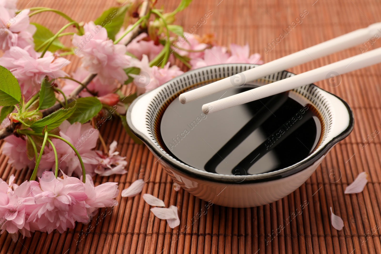 Photo of Bowl of soy sauce, chopsticks and beautiful blossoming branch on bamboo mat, closeup