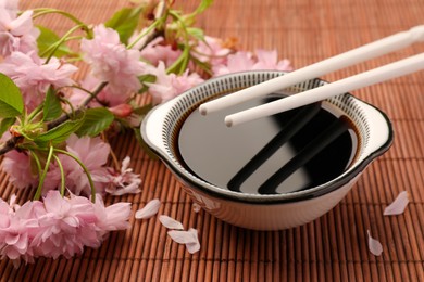 Bowl of soy sauce, chopsticks and beautiful blossoming branch on bamboo mat, closeup