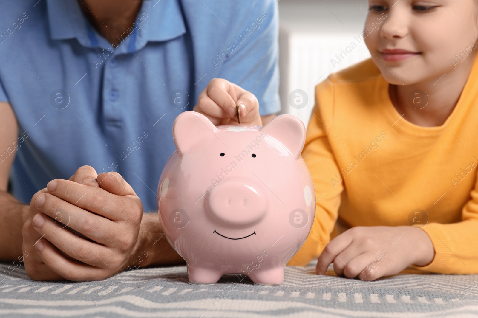 Photo of Little girl with her father putting coin into piggy bank at home, closeup