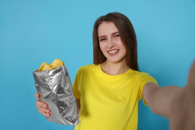 Pretty young woman with bag of tasty potato chips taking selfie on light blue background