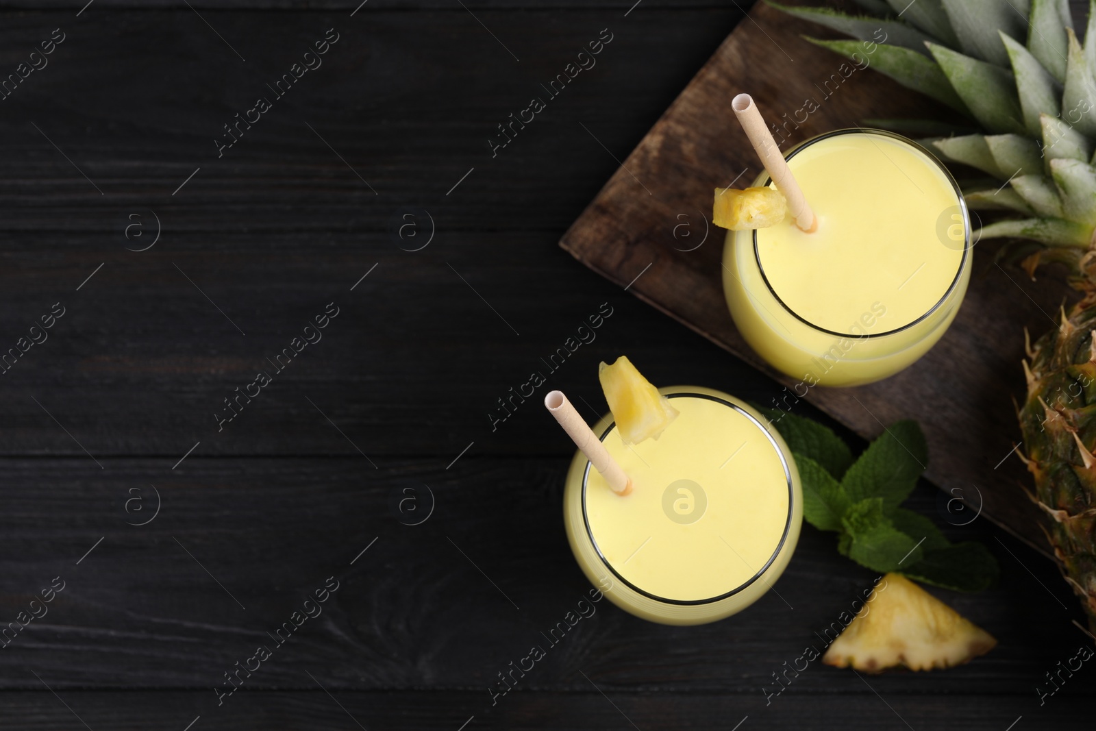 Photo of Tasty pineapple smoothie in glasses on black wooden table, flat lay. Space for text