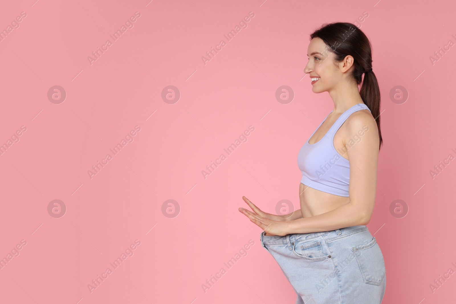 Photo of Happy young woman in big jeans showing her slim body on pink background, space for text