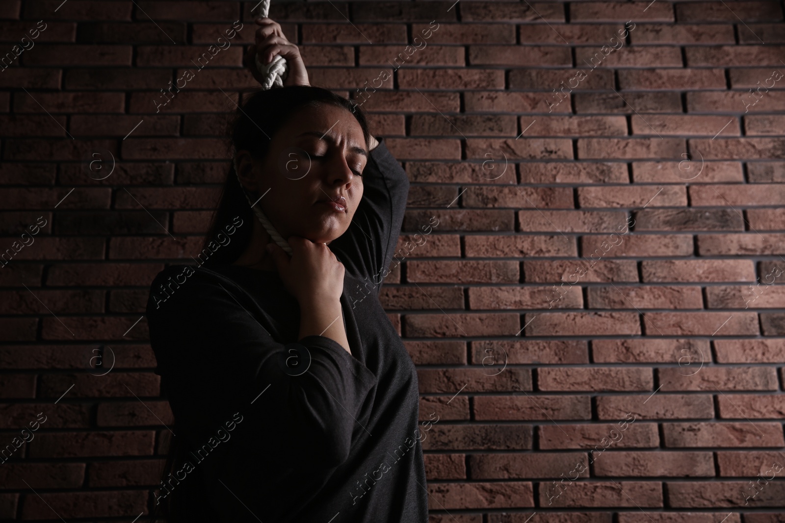 Photo of Depressed woman with rope noose on neck near brick wall. Space for text