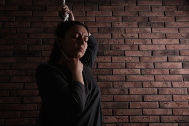 Photo of Depressed woman with rope noose on neck near brick wall. Space for text