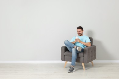 Happy man sitting in armchair and using smartphone indoors, space for text