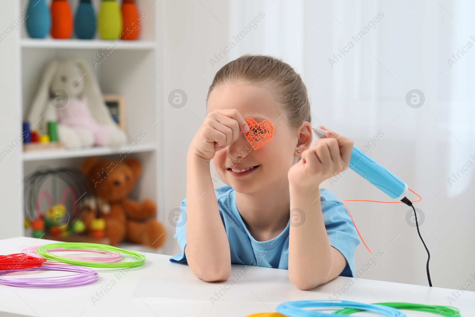 Photo of Girl holding heart made with stylish 3D pen near her eye at white table indoors