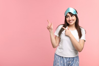 Happy woman in pyjama and sleep mask pointing at something on pink background, space for text
