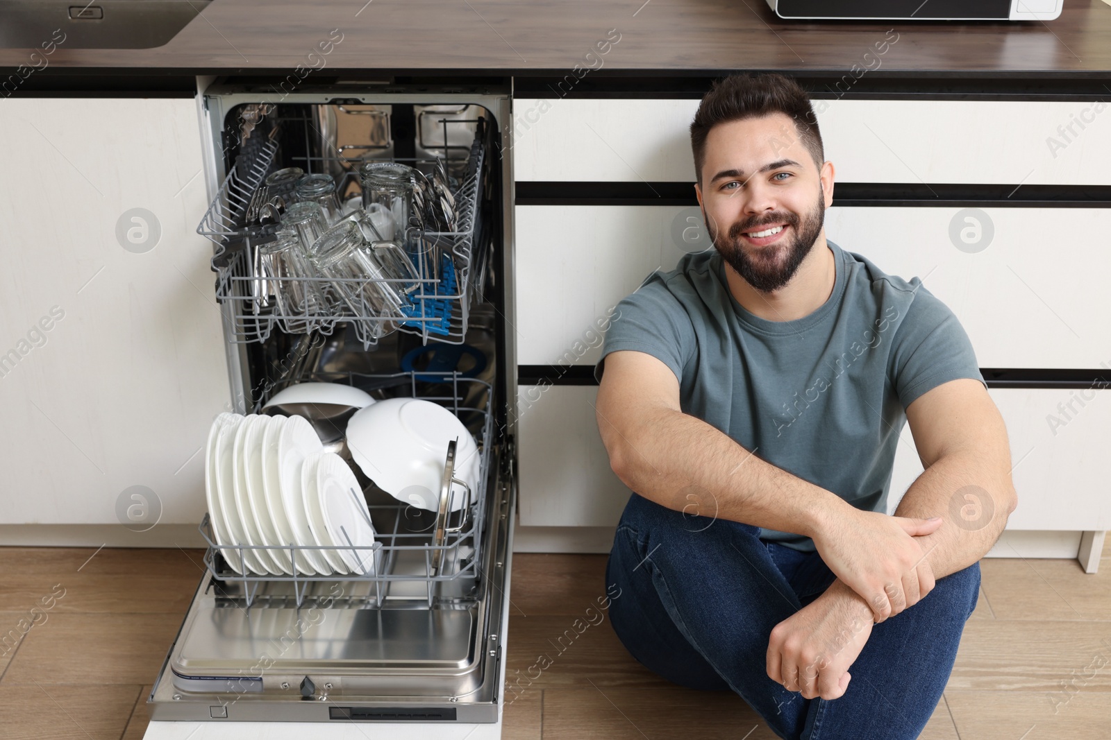 Photo of Smiling man sitting near open dishwasher in kitchen