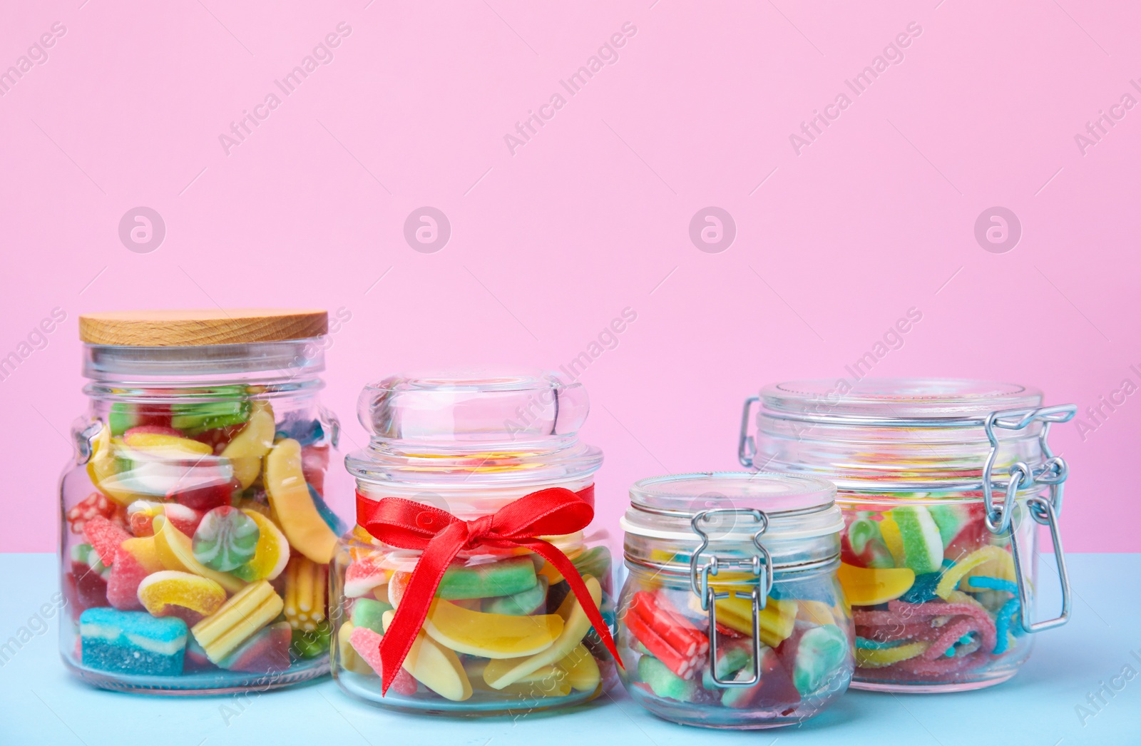 Photo of Tasty colorful jelly candies in glass jars on light blue table against pink background