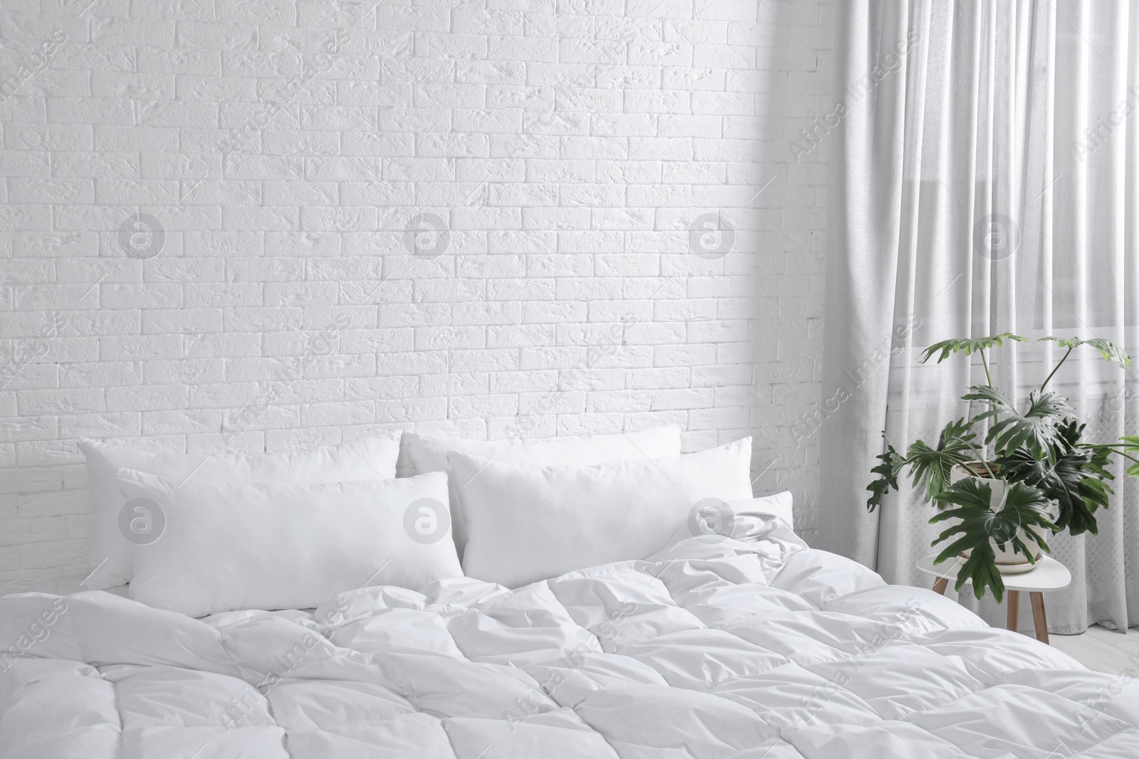 Photo of Large comfortable bed with pillows and blanket near white brick wall indoors. Stylish interior