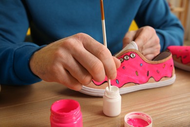 Photo of Man painting on sneaker at wooden table indoors, closeup. Customized shoes