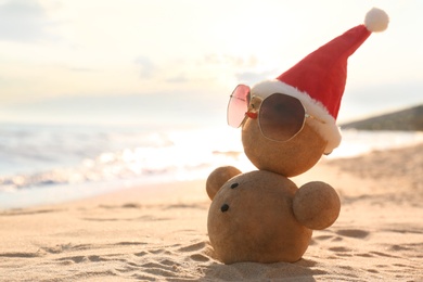 Snowman made of sand with Santa hat and sunglasses on beach near sea, space for text. Christmas vacation