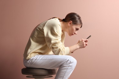 Photo of Woman with bad posture using smartphone while sitting on stool against pale pink background